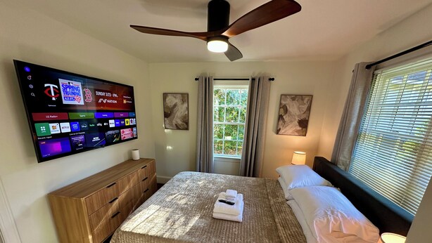 A hotel room with a bed, a TV mounted on the wall displaying various channels, a ceiling fan, and a window with curtains.