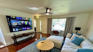 A living room with a flat-screen TV mounted on the wall, a wooden coffee table, a white sofa with blue cushions, and a patterned rug.