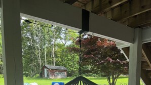 Un fauteuil suspendu avec des coussins, placé dans un espace extérieur abrité, d’où l’on peut admirer une cour verdoyante et un petit bâtiment.