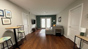 A living room with a wooden floor, a grey sofa, a small table, and a TV mounted on the wall. - Charming 2-bedroom apartment in Wilmington, NC (Wilmington)