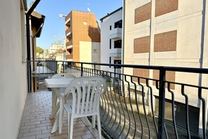 A balcony with a white table and chair, a metal railing, and buildings in the background.