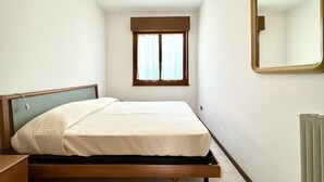 Ein kleines Zimmer mit einem Einzelbett, einem Holzkopfteil, einem Fenster mit Vorhängen und einem Spiegel an der Wand.