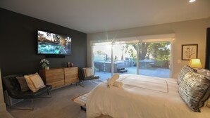 Ein modernes Schlafzimmer mit einem großen Bett, einem an der Wand montierten Fernseher, einer Couch und Blick ins Freie durch eine Schiebetür.