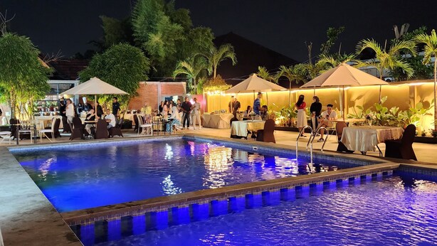 Un espace piscine aménagé pour la nuit, avec un bord carrelé, entouré de tables et de chaises placées sous des parasols, et quelques personnes réunies aux alentours.