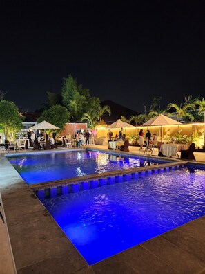 Un espace piscine aménagé pour la nuit, avec un bord carrelé, entouré de tables et de chaises placées sous des parasols, et quelques personnes réunies aux alentours.