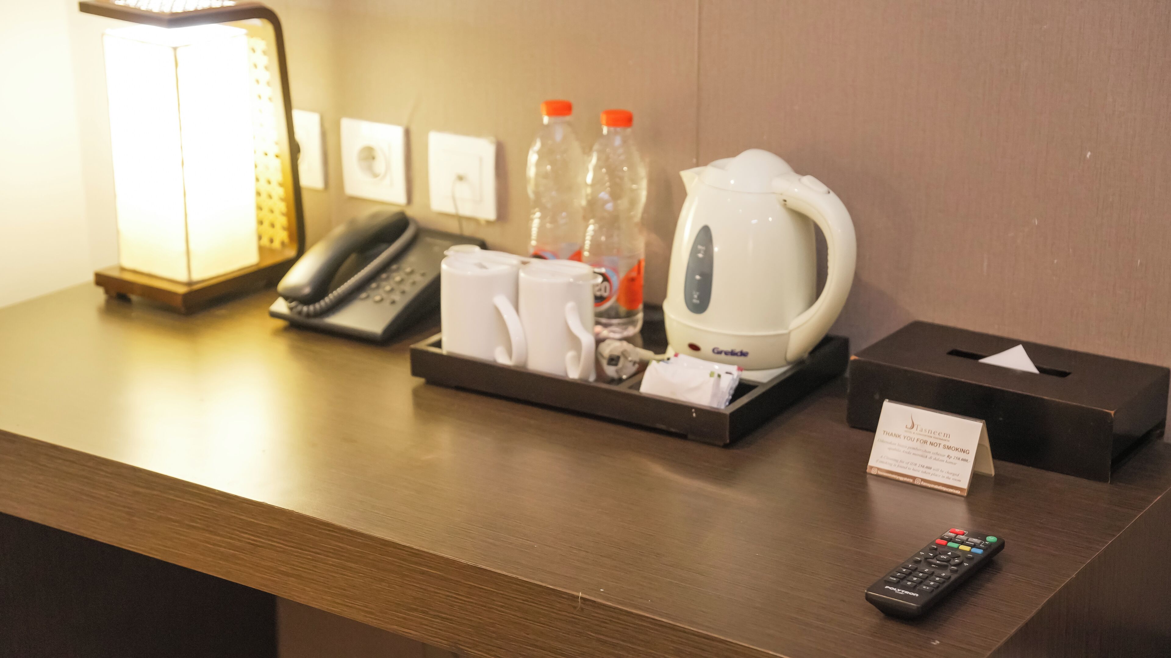 A hotel reception desk with a telephone, two bottles of water, a kettle, a tray with mugs, a remote control, and a no-smoking sign.