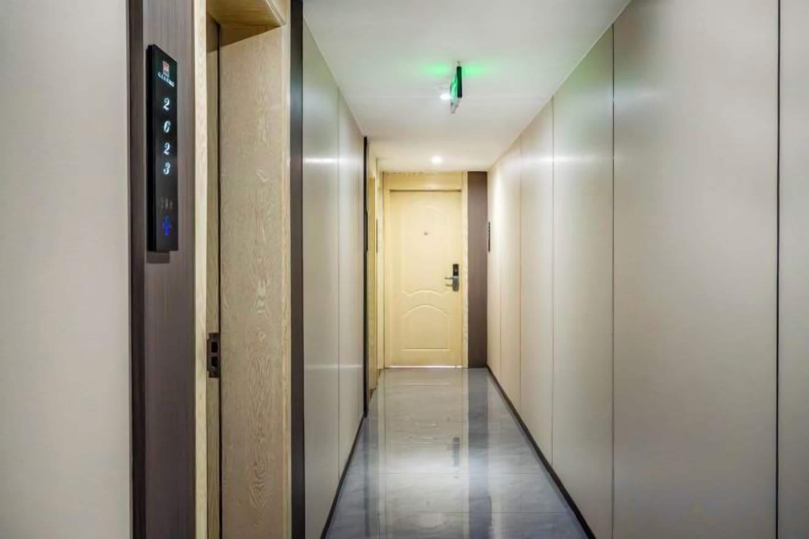 A hallway with a door at the end, a wall-mounted elevator control panel, and a light fixture on the ceiling.