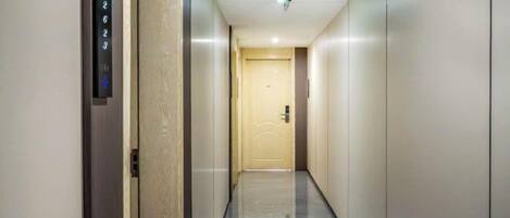 A hallway with a door at the end, a wall-mounted elevator control panel, and a light fixture on the ceiling.