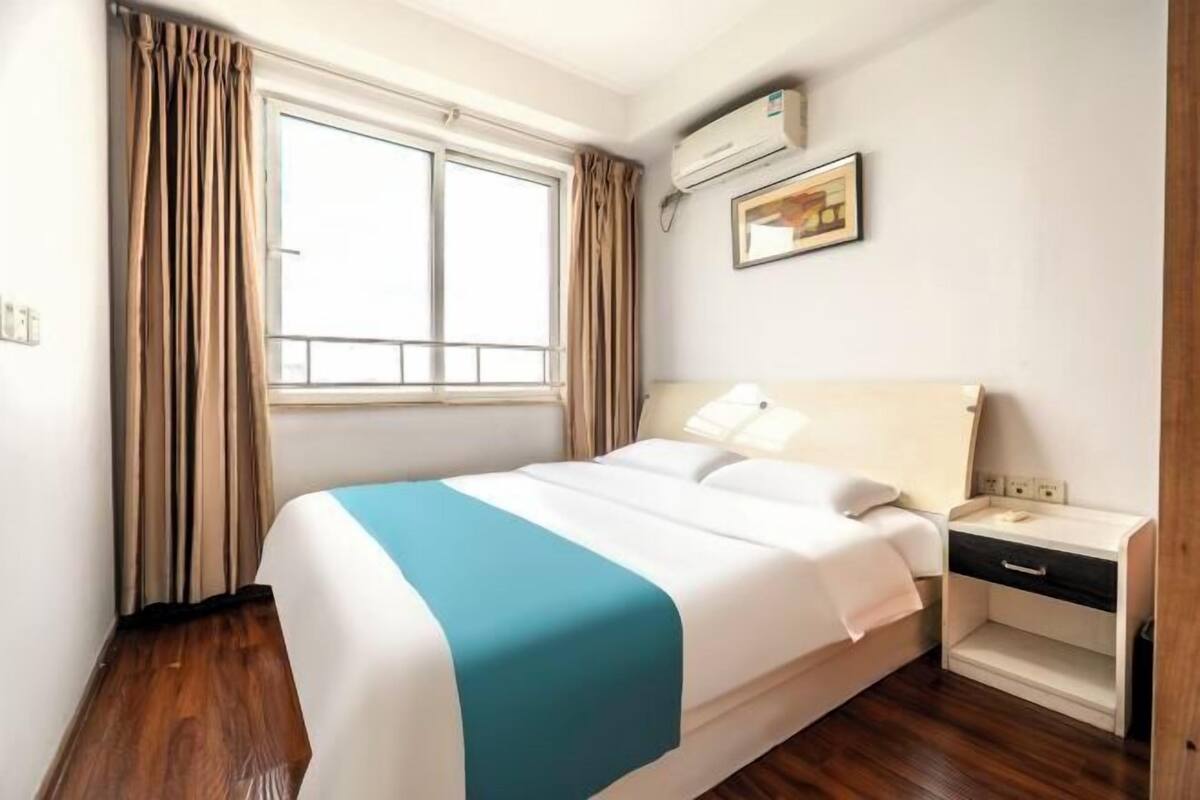A hotel room with a bed, a nightstand, a window with curtains, an air conditioning unit, and a framed picture on the wall.