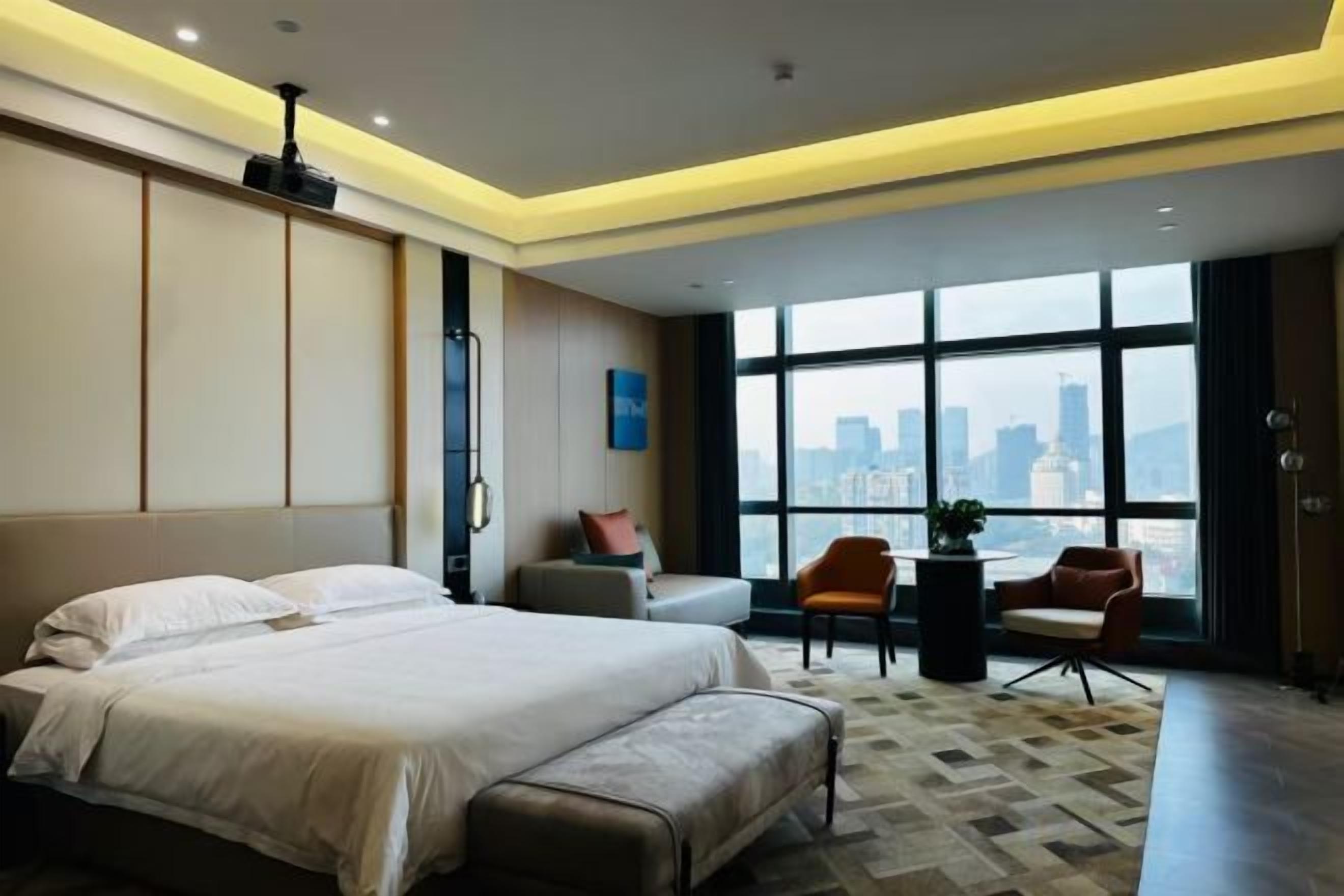 A modern hotel room with a large bed, a seating area with a table and chairs, and a view of a cityscape through large windows.