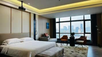 A modern hotel room with a large bed, a seating area with a table and chairs, and a view of a cityscape through large windows.