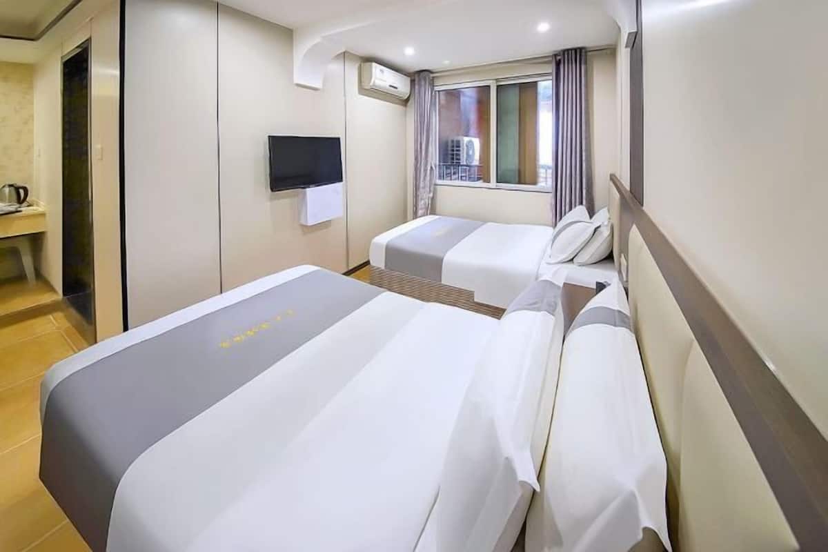 A hotel room with two beds, a TV, an air conditioning unit, and a window with curtains.