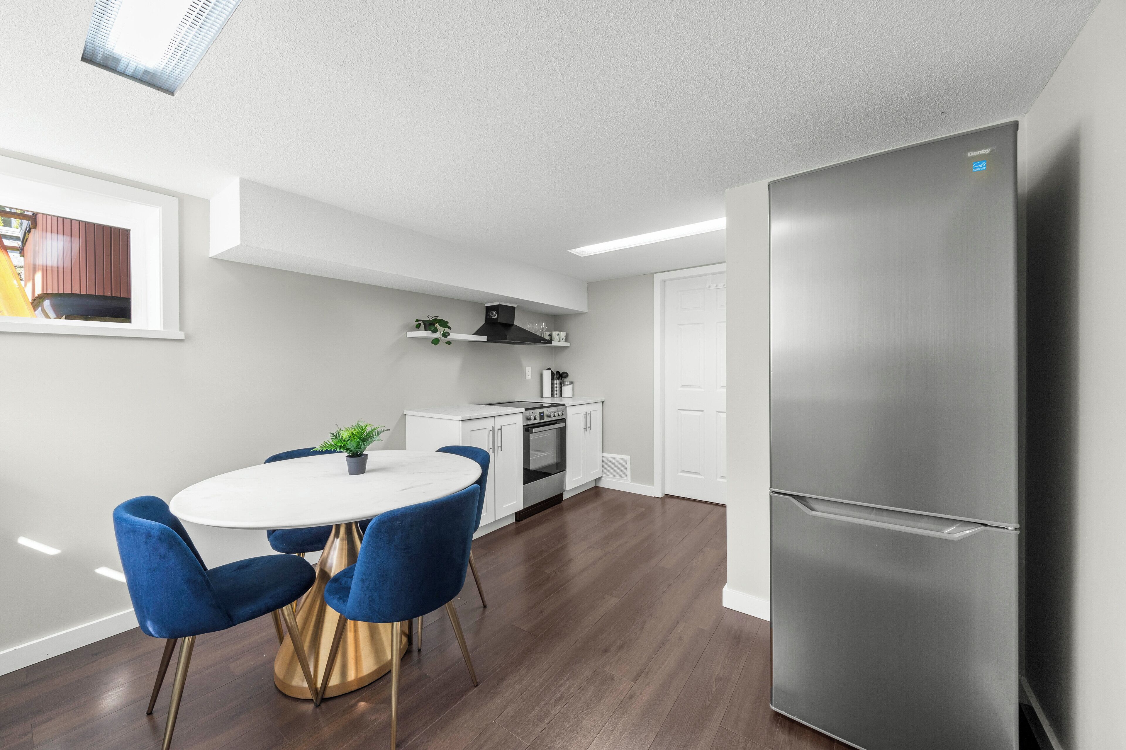 A modern kitchen with a stainless steel refrigerator, white cabinetry, and a round dining table with blue chairs.
