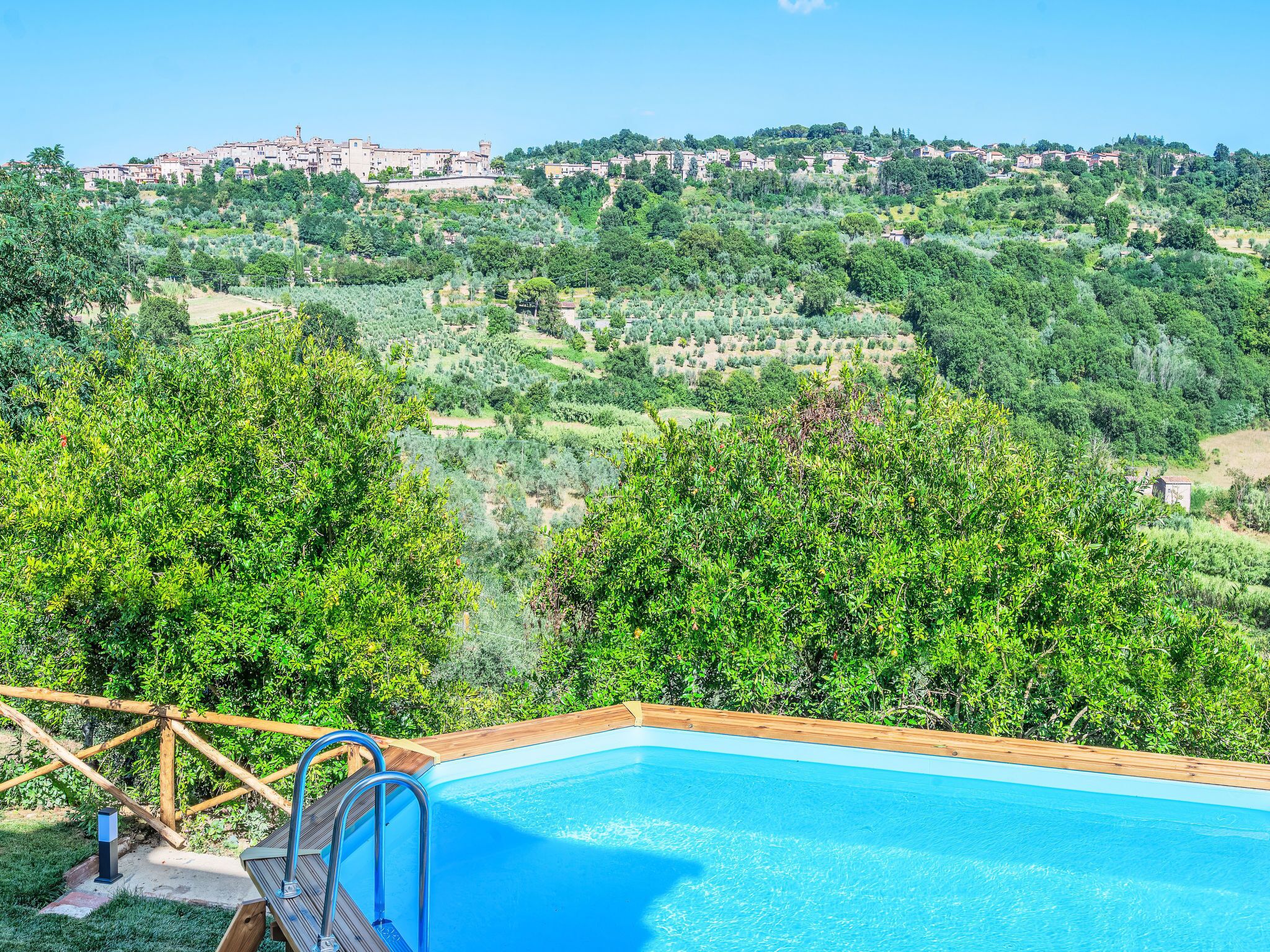 A swimming pool with a wooden deck and metal ladder, surrounded by lush greenery and overlooking a hillside with buildings.