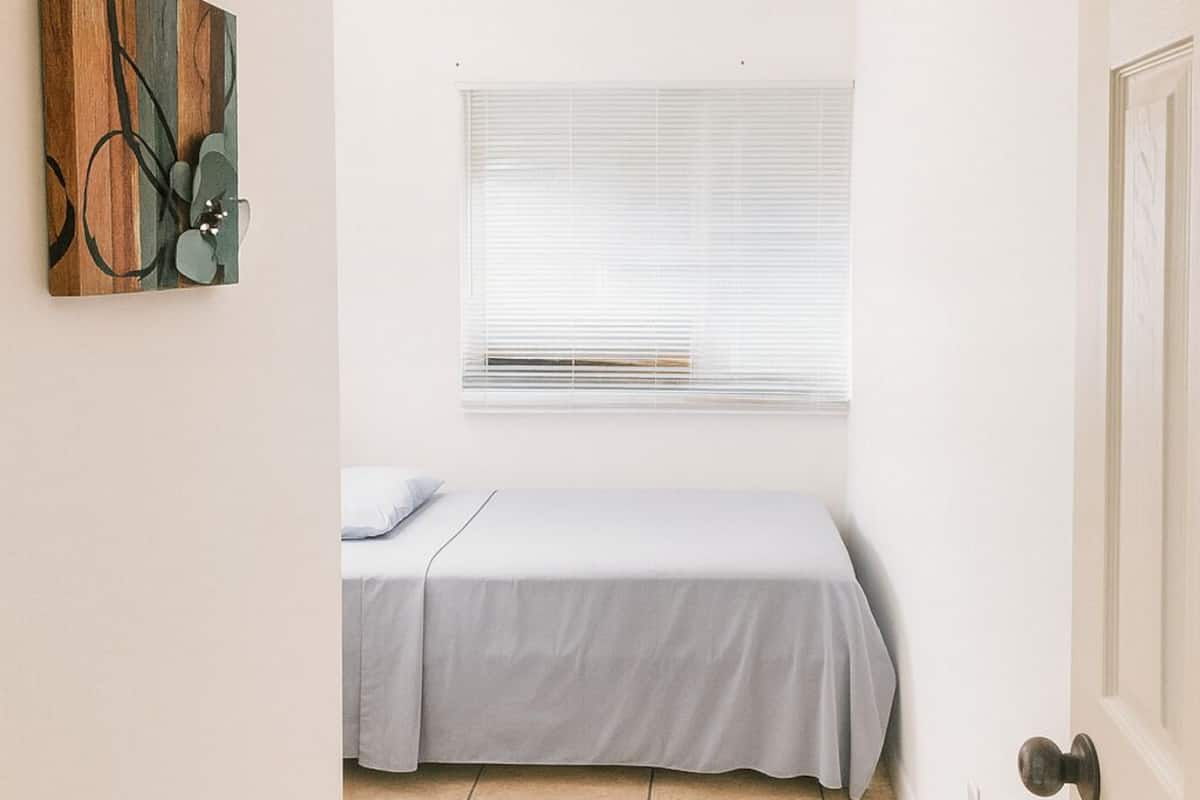 A small, narrow room with a bed, a window with blinds, a door, and a wall-mounted abstract art piece.