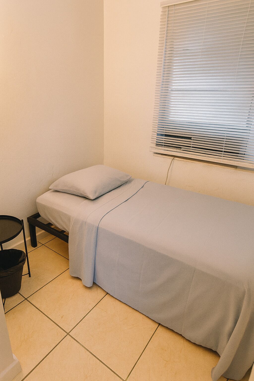 Ein kleines Zimmer mit einem Einzelbett, einer hellen Matratze, einem Kissen, einem kleinen Tisch und einem Fenster mit Jalousien.