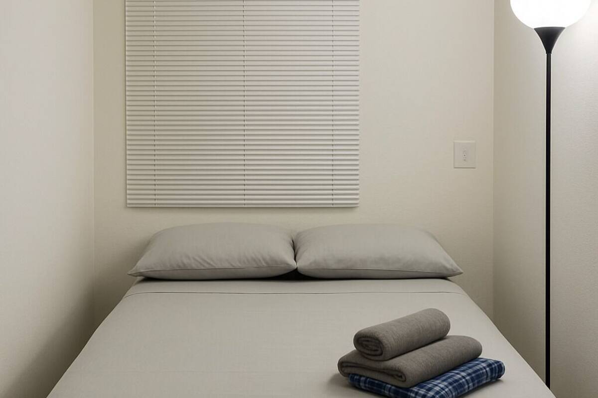 A bed with a light-colored bedspread, two pillows, and a folded blanket. There is a floor lamp to the right and a window with blinds above the bed.