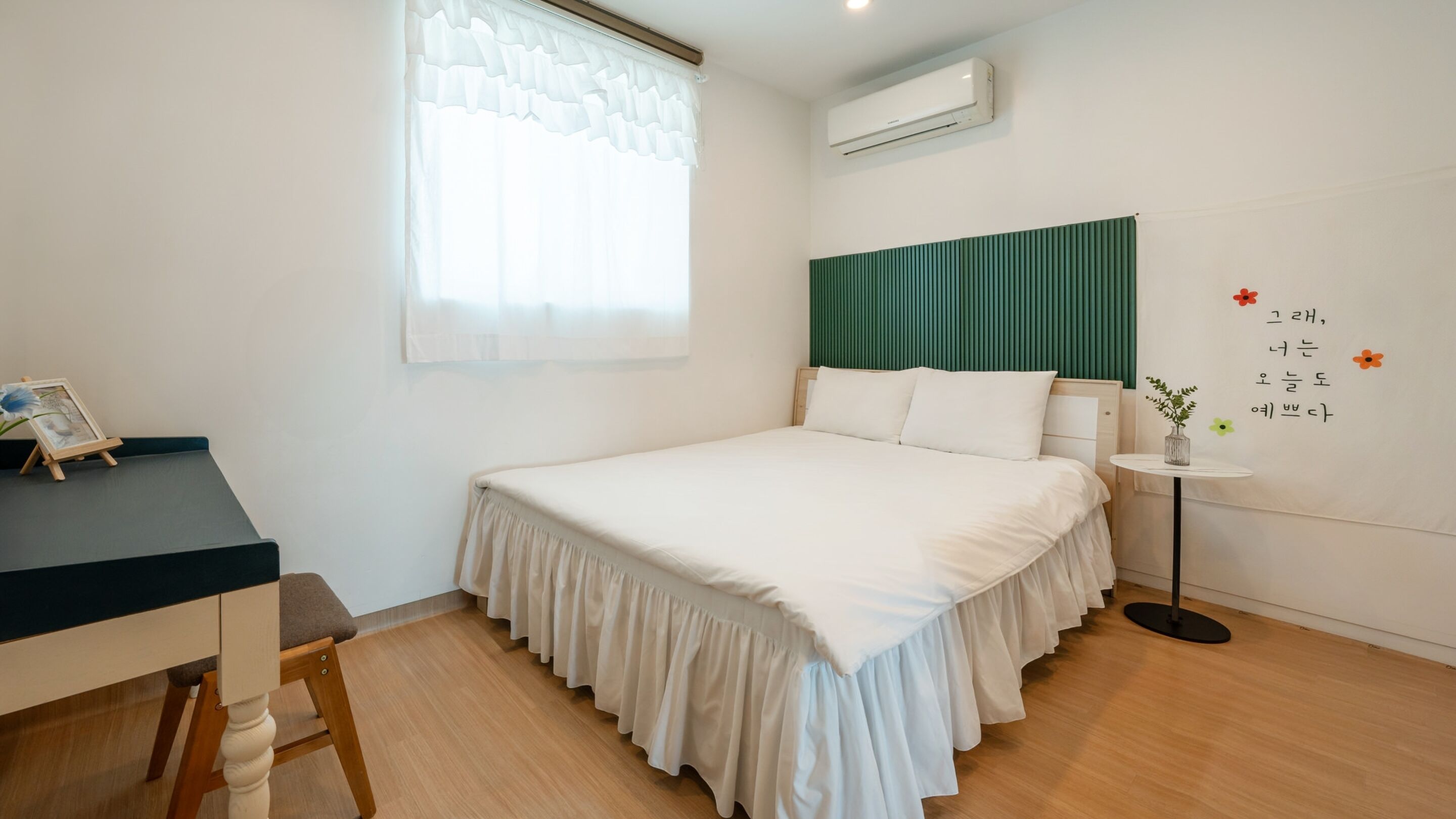 A bedroom with a bed, a desk, a chair, a window with curtains, an air conditioner, and a wall with a decorative item.