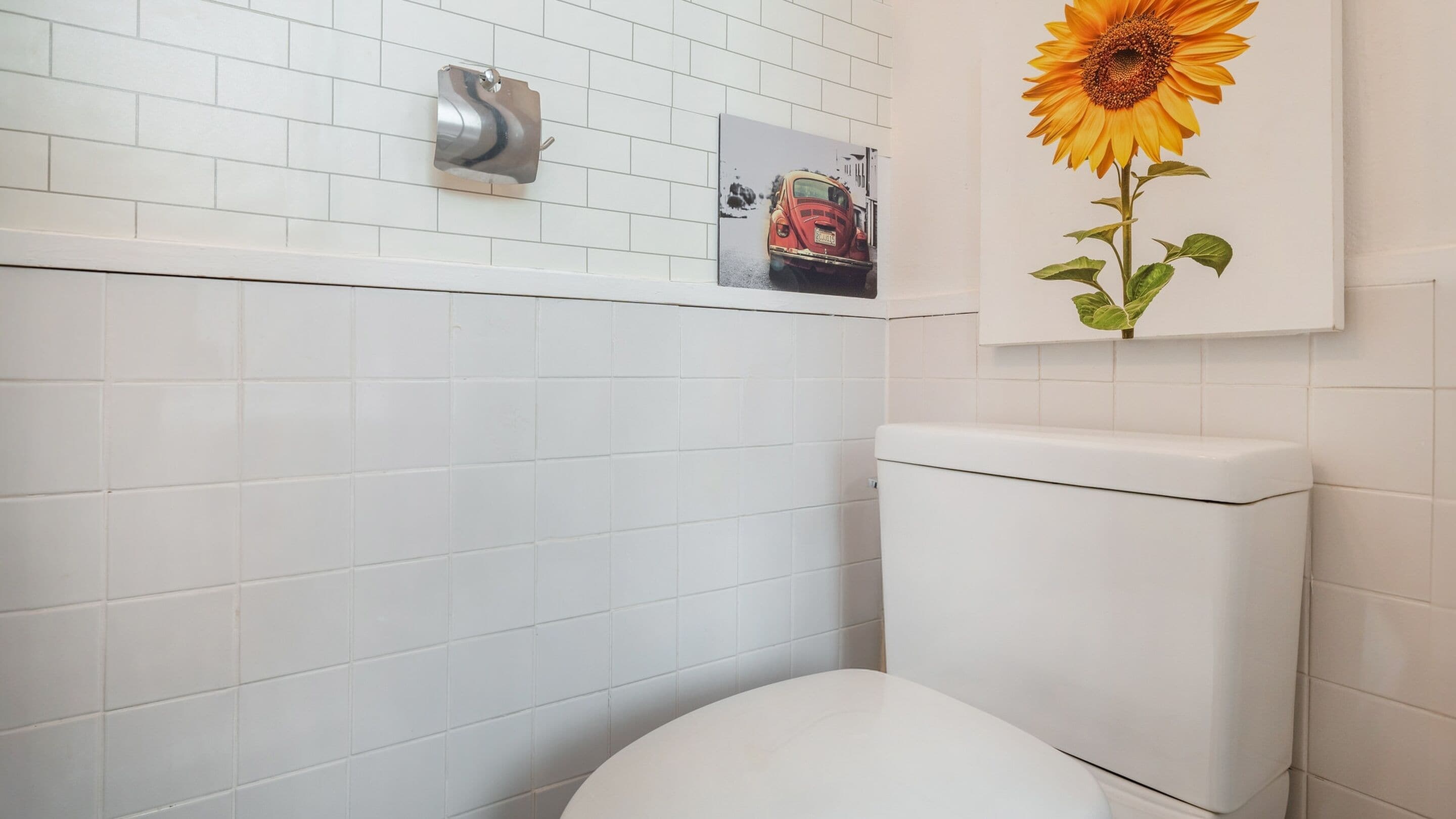 Een badkamer met witte betegelde wanden, een wit toilet, een handdroger en een lijst met een zonnebloem.