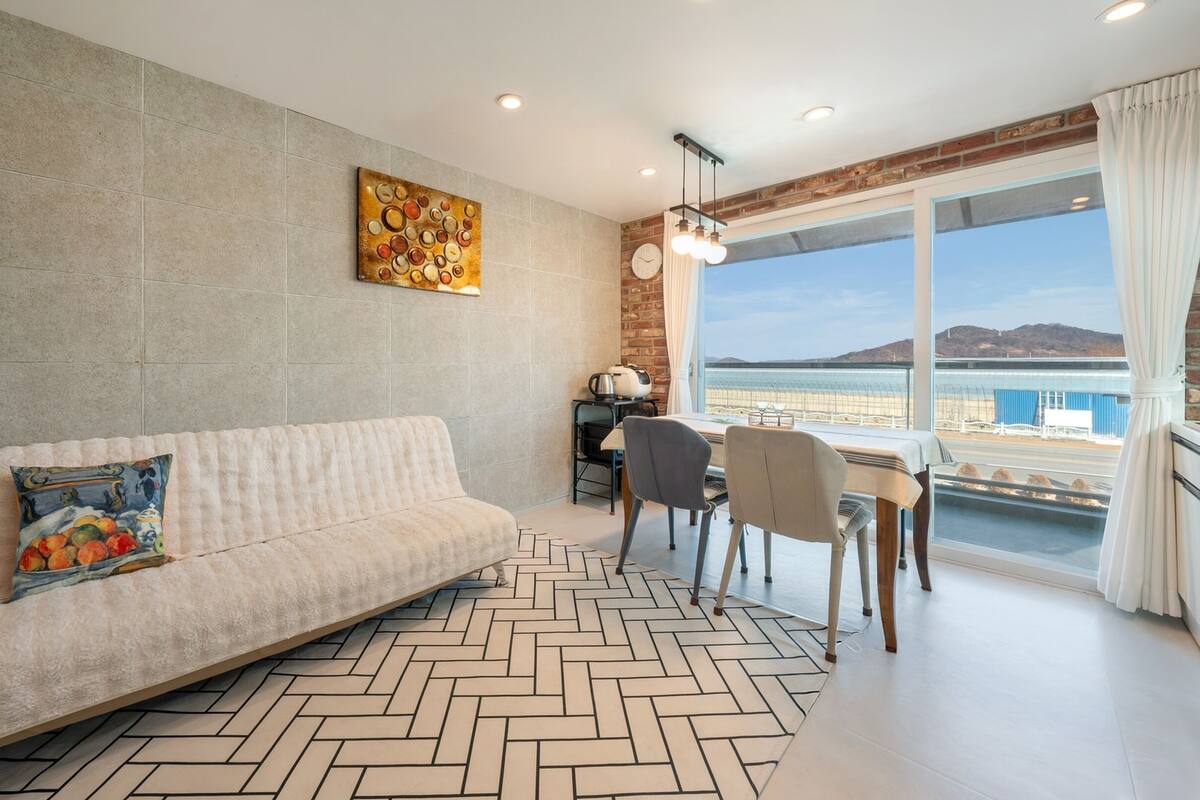 A modern living room with a sofa, a dining table with chairs, a kitchen area, and a large window with a view of the sea.