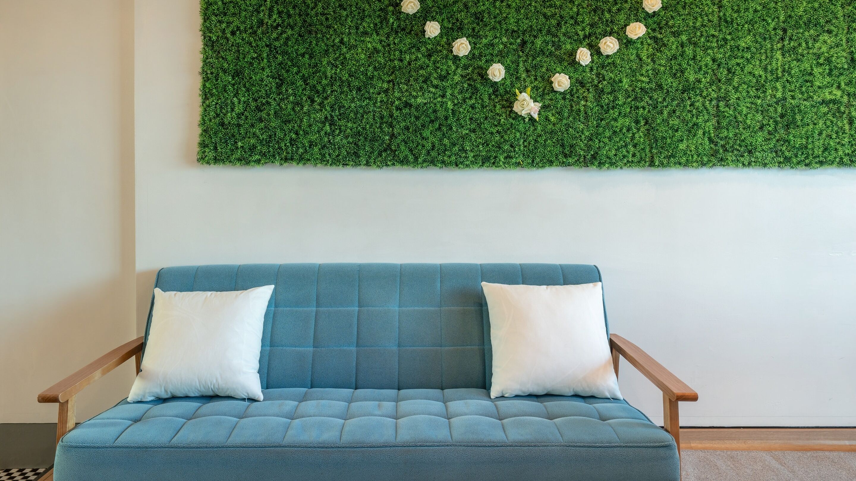Un sofá azul con dos cojines blancos frente a un muro con un fondo verde texturizado y flores blancas.