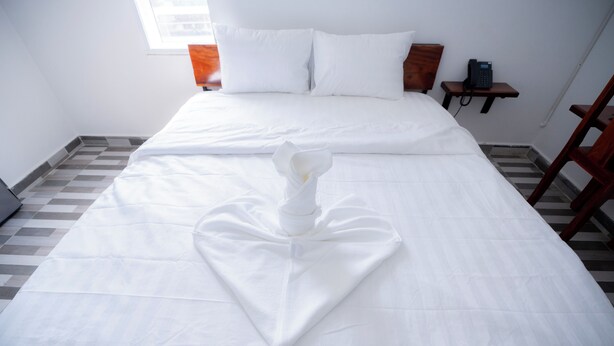 A neatly made bed with a heart-shaped towel in the center, a bedside table with a phone, and a window with blinds. - Bona Hostel PP (Phnom Penh)