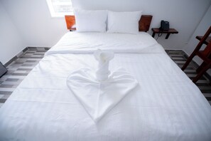 A neatly made bed with a heart-shaped towel in the center, a bedside table with a phone, and a window with blinds. - Bona Hostel PP (Phnom Penh)