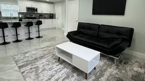 Un salon moderne avec un canapé en cuir noir, une table basse blanche et une cuisine en arrière-plan.
