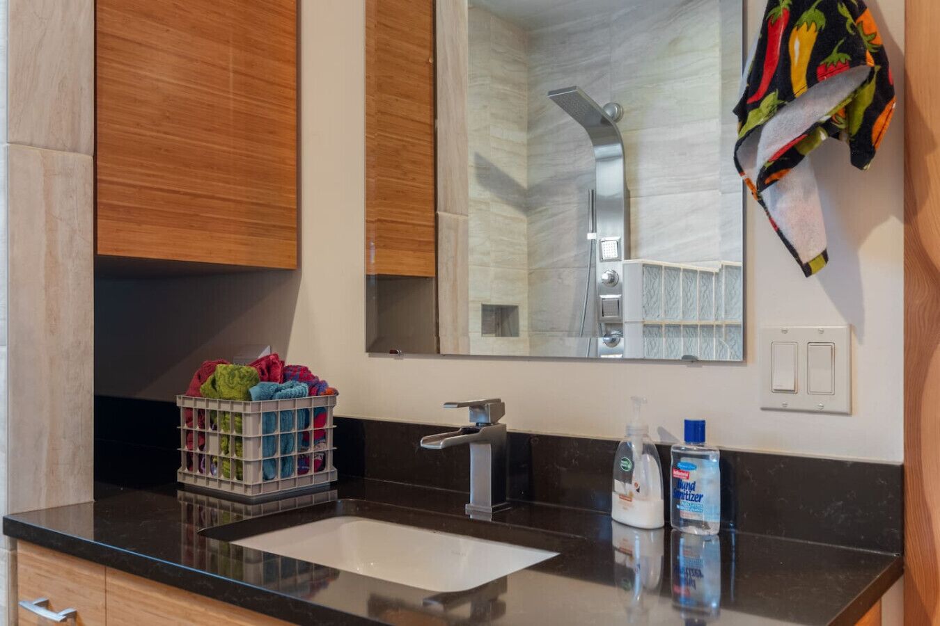 A bathroom with a black countertop, a white sink, a mirror, and a shower with a chrome head. There are towels hanging on the wall and a basket of colorful towels on the counter.