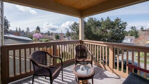 A covered outdoor deck with two wicker chairs and a small table, overlooking a residential area with houses and trees. - Custom Charm! Central - Office & Peaceful yard! (Seattle)