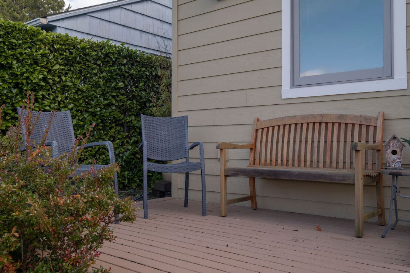 Un banc en bois et deux chaises en osier sont placés sur une terrasse en bois, avec une haie et une maison en arrière-plan.