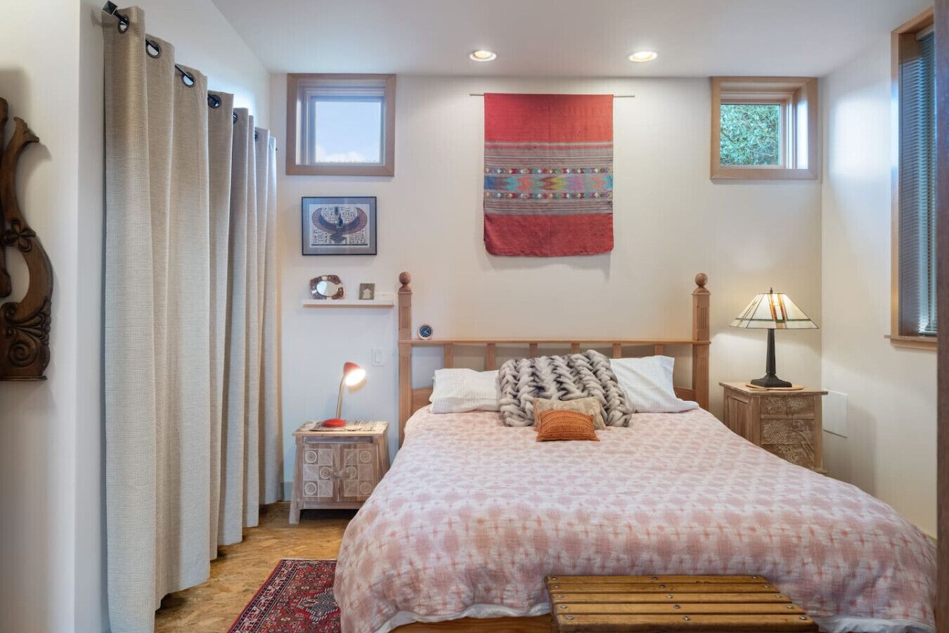 Ein Schlafzimmer mit Bett, Nachttischen, einer Lampe, einem roten Wandteppich und einem kleinen Fenster.