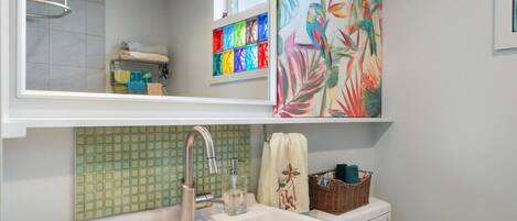 Un baño con un lavabo blanco, salpicadero de azulejos verdes, un espejo, un perchero y un cuadro abstracto colorido.
