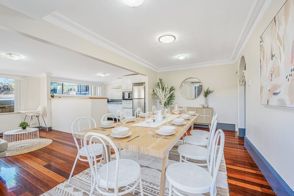 Un espace repas avec une longue table en bois, dressée pour six personnes, entourée de chaises blanches, dans une pièce au plancher et aux murs blancs.