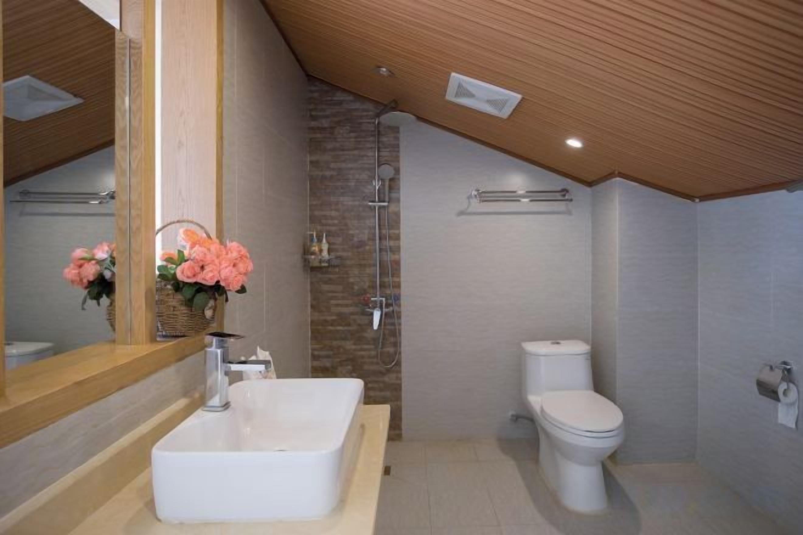 A modern bathroom with a wooden ceiling, a large mirror, a white sink, a shower, and a toilet.