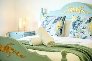 Un lit avec une couverture verte tricotée, une serviette blanche roulée et un oreiller orné d’un motif de feuilles tropicales.