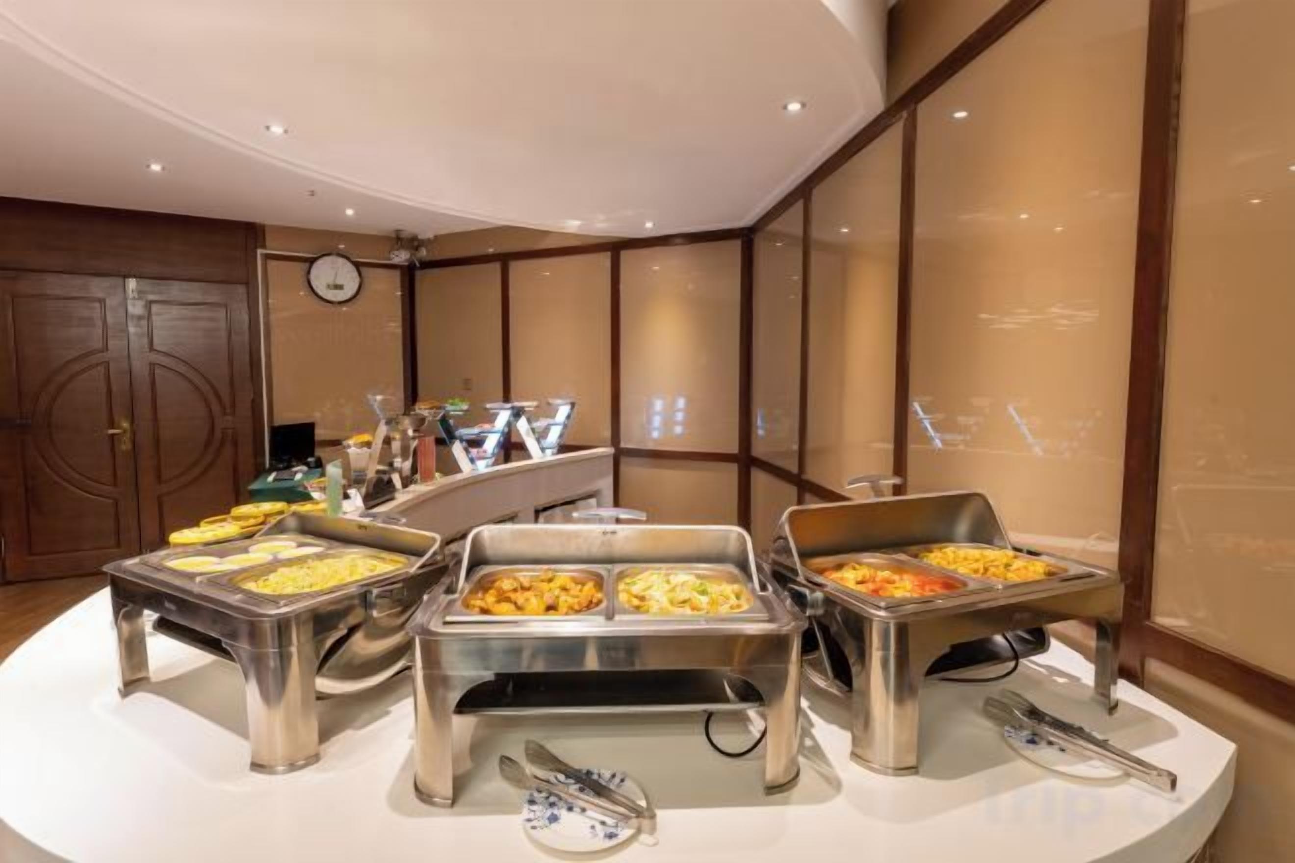 Disposición de buffet con múltiples dispensadores de alimentos, un reloj en la pared y una puerta de madera al fondo.