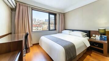 A hotel room with a bed, a desk, a chair, a nightstand with a lamp, and a window with curtains.