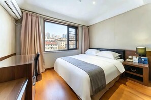 A hotel room with a bed, a desk, a chair, a nightstand with a lamp, and a window with curtains. - Regal Hotel (Jiamusi Railway Station Department Store) (Jiamusi)