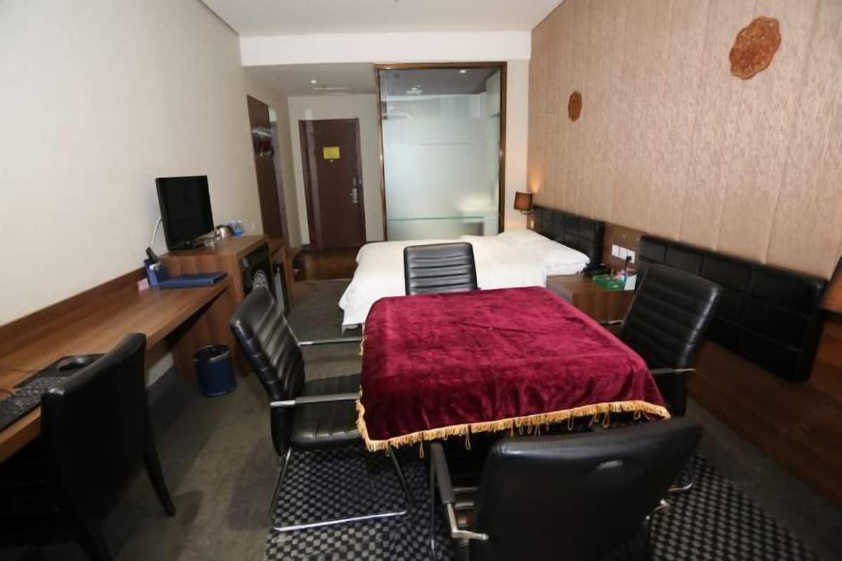 A hotel room with a bed, a desk with a computer, a chair, a TV, and a bathroom visible through a glass door.