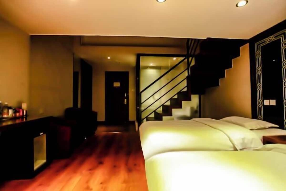 A modern bedroom with a wooden floor, a bed with white linens, a desk with a mirror, and a staircase leading to an upper level.