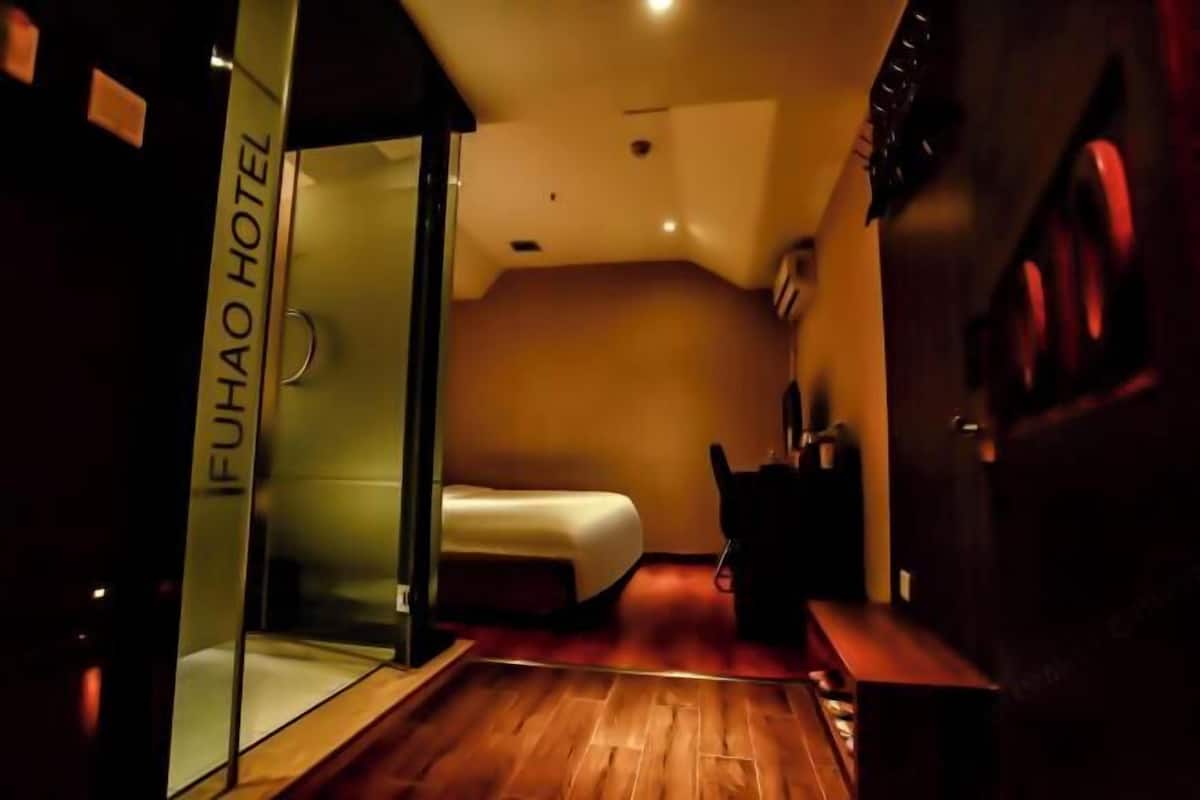 A hotel room with a bed, a glass partition, a wooden floor, and a bench.