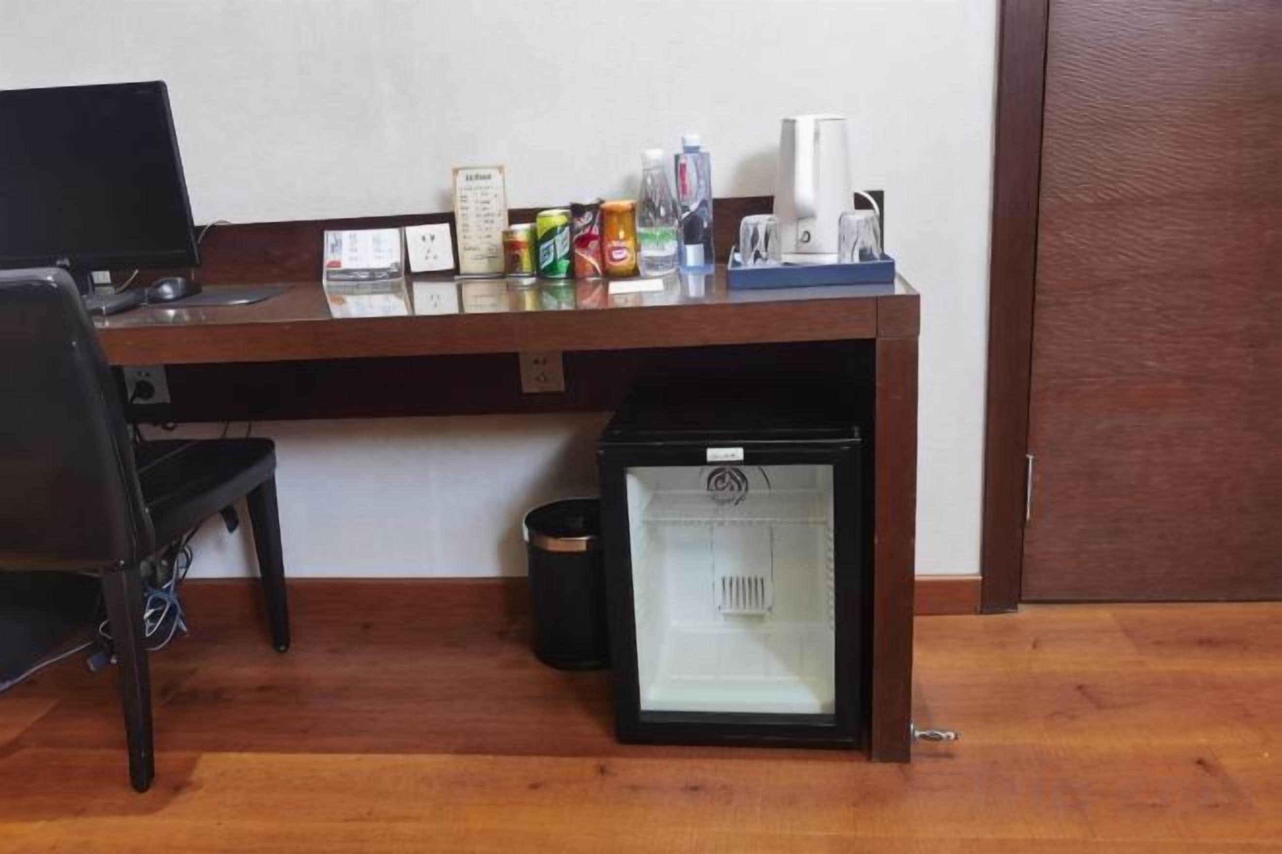 A room with a wooden desk, a computer, a small refrigerator, and a trash bin.