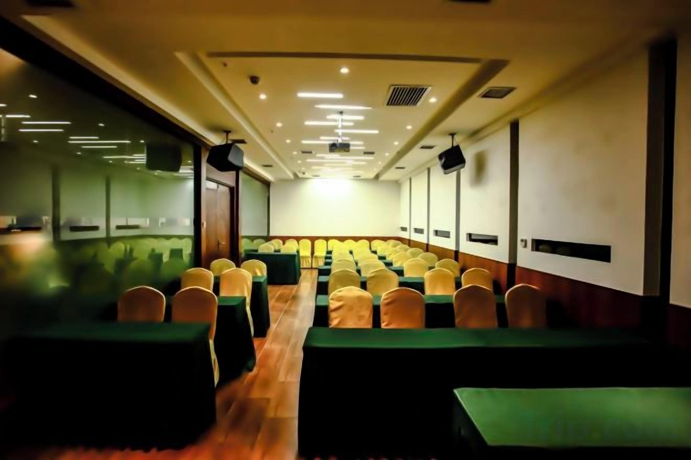 Un salón de conferencias con filas de mesas y sillas, una pantalla de proyector y una zona de escenario.