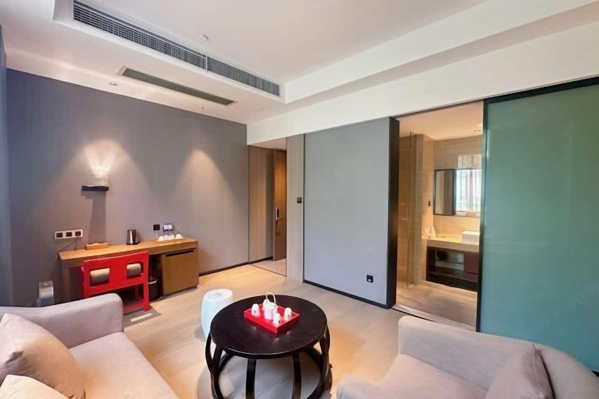 Quarto de hotel moderno com sofá, uma pequena mesa redonda, escrivaninha com cadeira e porta deslizante para o banheiro.