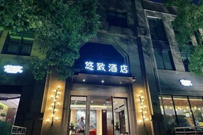 Entrada de hotel con señalización iluminada, fachada de vidrio y un árbol en primer plano.