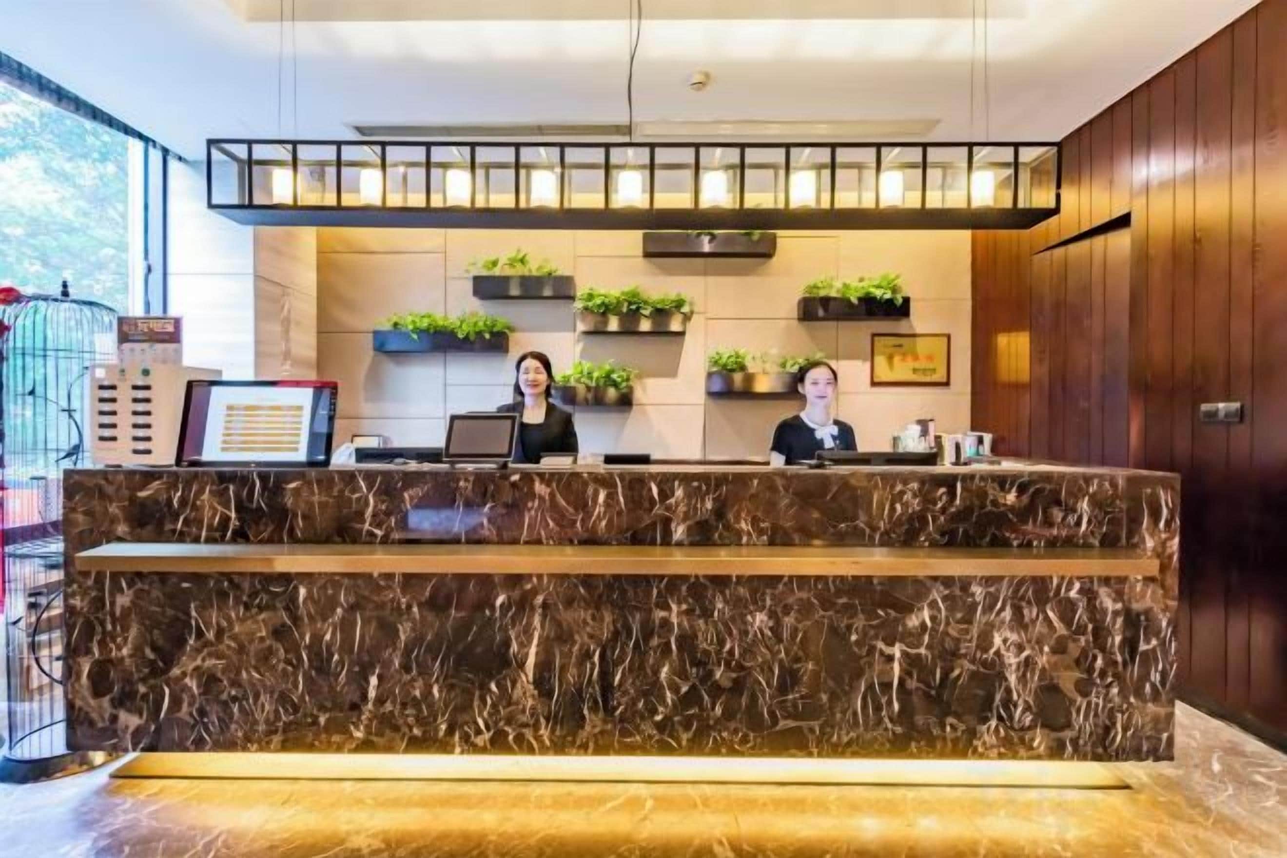 A reception desk with two staff members, a marble counter, and potted plants on shelves.
