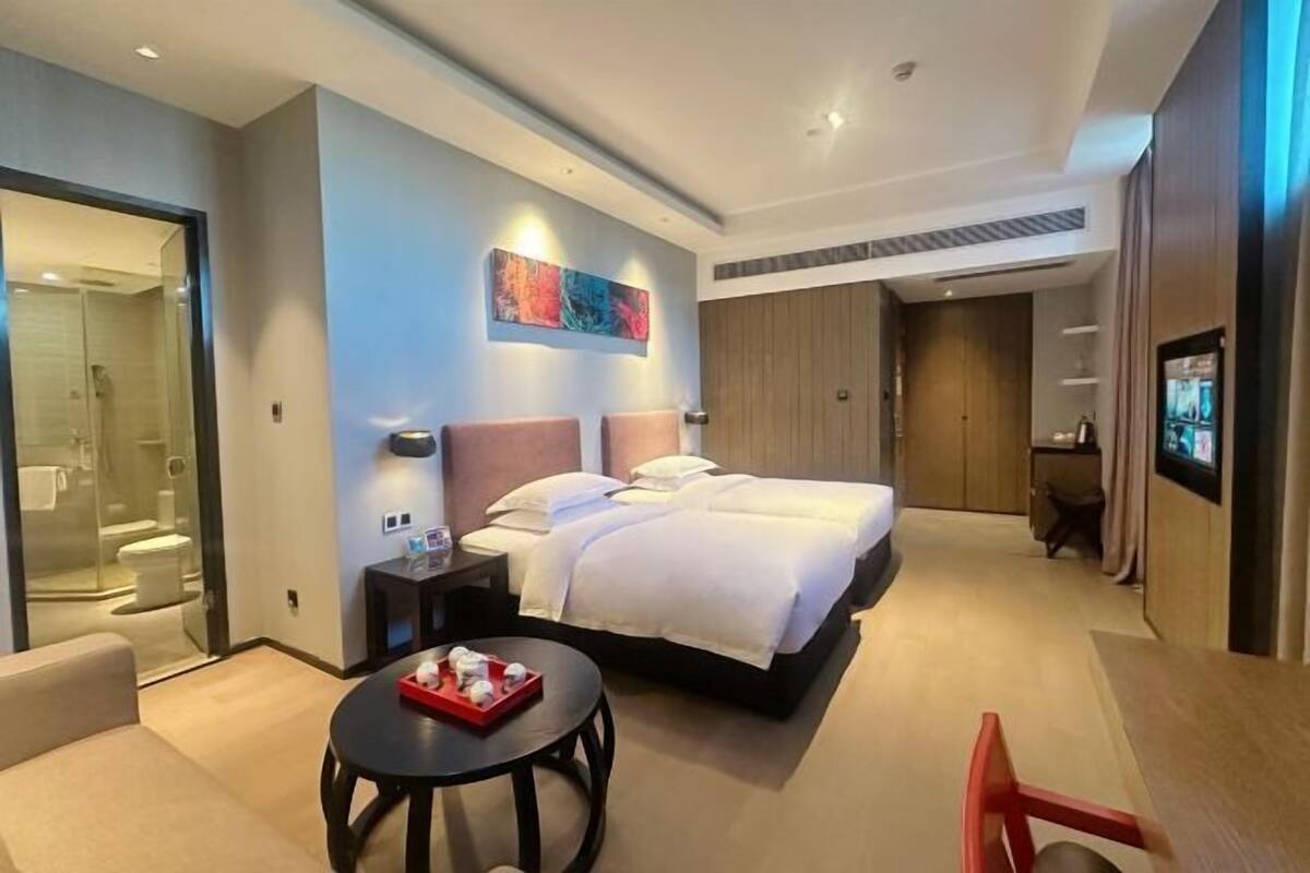 Quarto de hotel moderno com uma cama grande, uma pequena mesa redonda com um tabuleiro de produtos de higiene pessoal, uma TV fixada na parede e uma área de chuveiro com divisória de vidro.