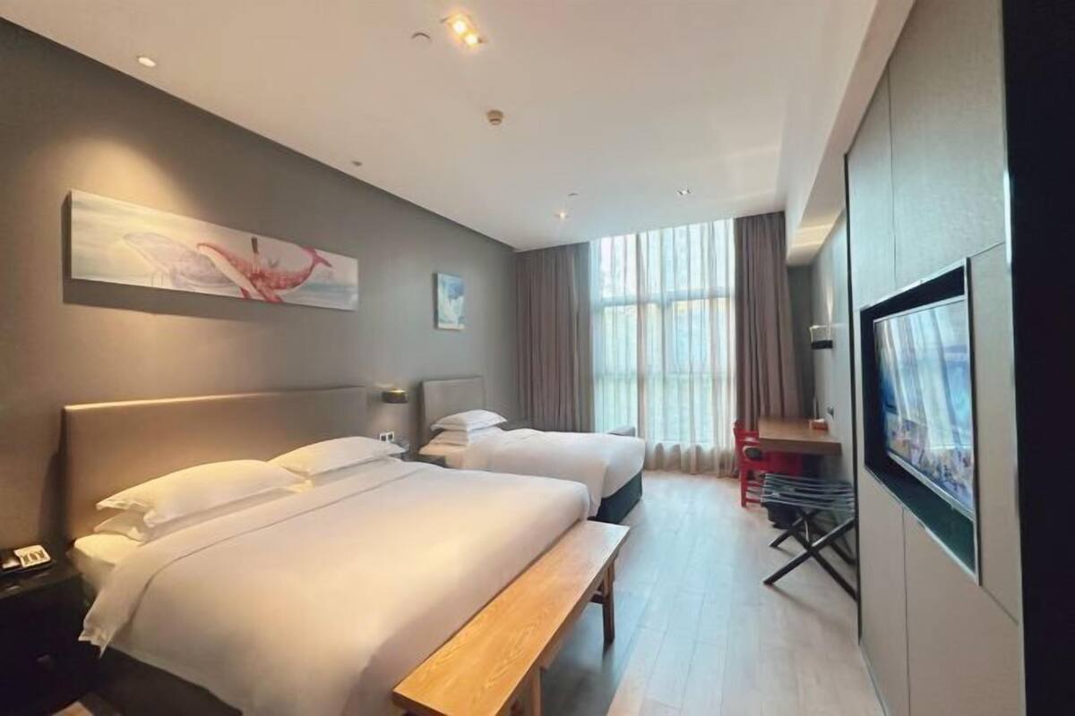 Quarto de hotel com duas camas, um banco, uma TV e uma janela com cortinas.
