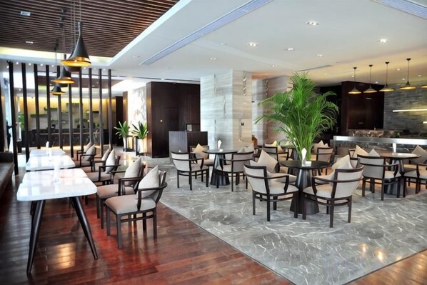 A modern hotel lobby with multiple seating areas, including tables and chairs, and a large plant in the center.